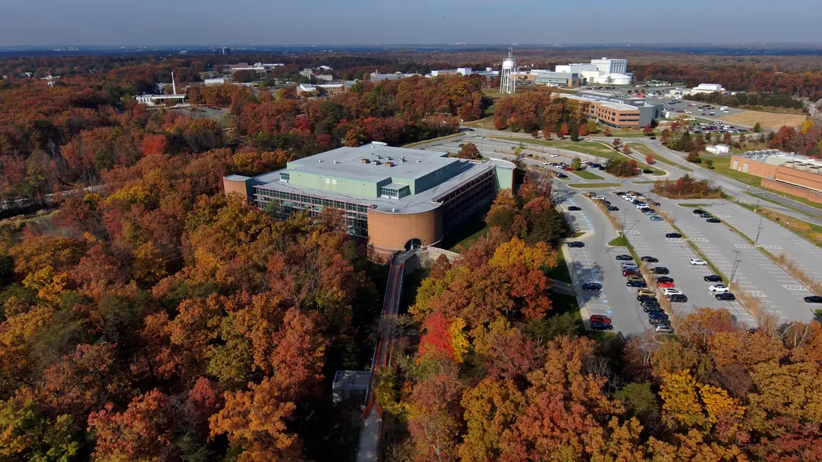 Aerial view of a large commercial building with a flat roof beside a parking lot, surrounded by autumn woodland