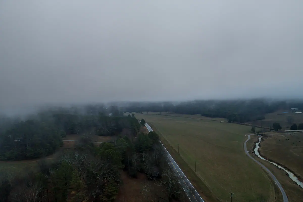 Aerial view of a rural road, tree line and creek with low fog over open land